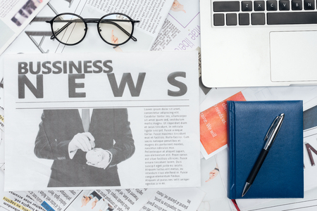 elevated view of eyeglasses, pen and notebook and laptop on newspapersの写真素材
