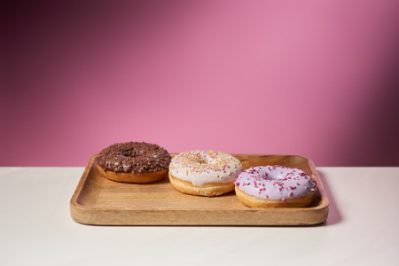 tasty donuts with icing on wooden cutting board on pink background