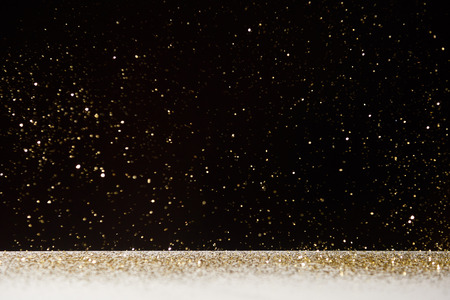 selective focus of golden sparkles falling on white table isolated on blackの写真素材