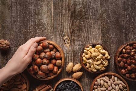 partial view of woman holding bowl with hazelnuts on wooden tabletop with different nuts aroundの写真素材