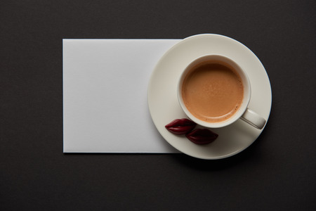 top view of cup with coffee, chocolate lips on saucer and empty white card on black backgroundの写真素材