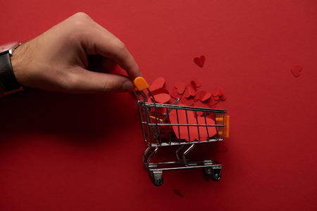 cropped view of male hand and toy shopping cart with paper cut hearts on red backgroundの写真素材