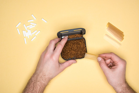 Cropped view of man with snuffbox making cigarettes isolated on yellow
