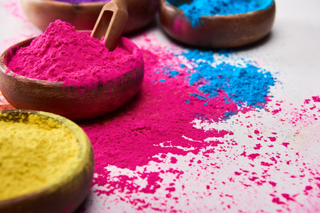 Wooden spatula and bowls with pink, blue and yellow holi powder on white backgroundの写真素材