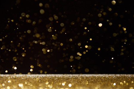 selective focus of golden shiny sparkles falling on table isolated on blackの写真素材