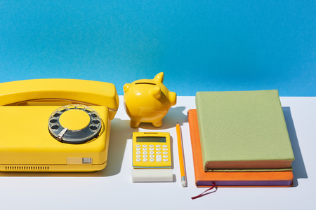 multicolored notebooks, calculator, telephone and piggy bank on white desk and blue backgroundの写真素材