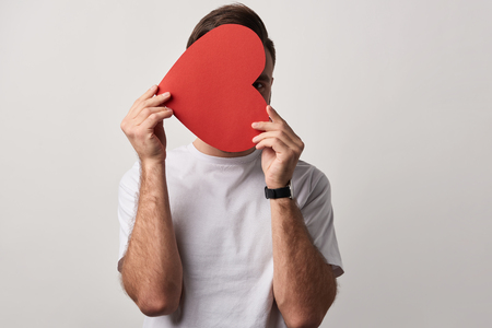 man hiding face behind empty red paper cut heart card isolated on greyの写真素材