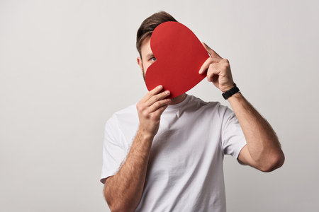 man hiding face behind blank paper cut heart card isolated on greyの写真素材