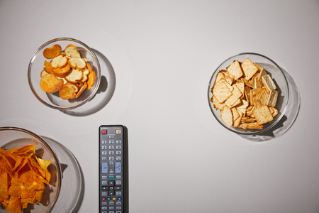 top view of glass bowls with tasty snacks near remote control on white backgroundの写真素材