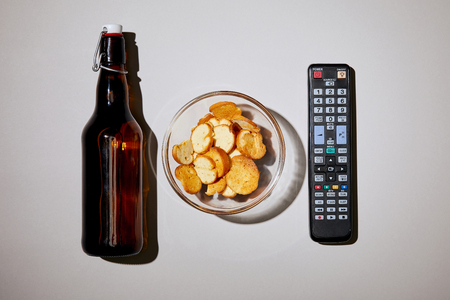 top view of brown bottle with beer near snack in bowl and remote control on white backgroundの写真素材