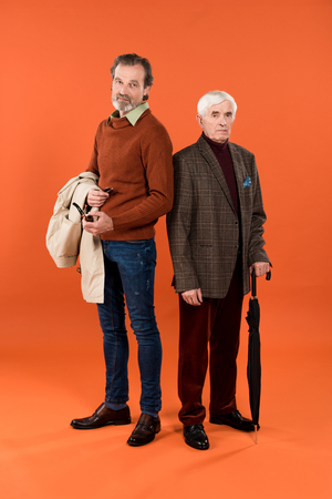 trendy retired man standing with jacket in hands near friend with umbrella on orange backgroundの写真素材