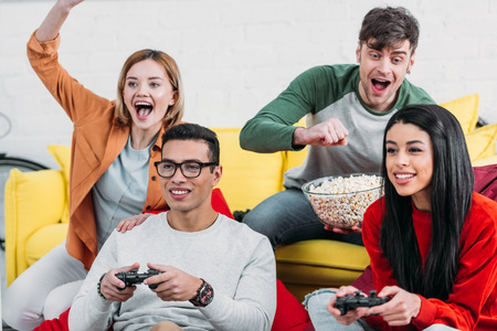 multicultural friends having fun at home party, playing video game and enjoying drinks and snacks