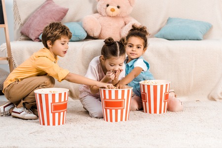 cute multiethnic children sitting on carpet and eating popcornの写真素材