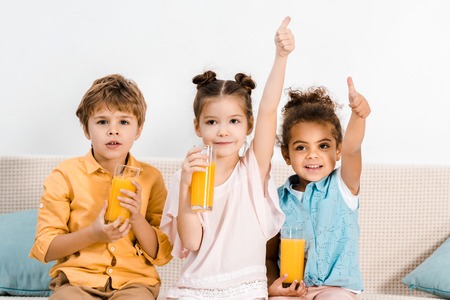 adorable multiethnic children holding glasses of juice and showing thumbs upの写真素材