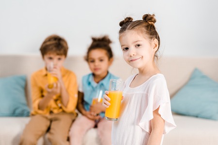 adorable child holding glass of juice and smiling at camera while little friends drinking juice behindの写真素材
