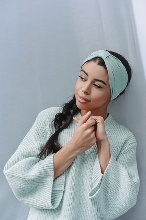 attractive mixed race young woman in turquoise sweater and headband at home and looking awayの写真素材