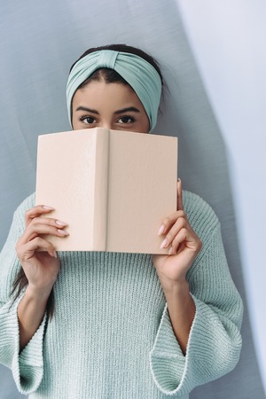 beautiful mixed race girl in turquoise sweater and headband looking out from book at home