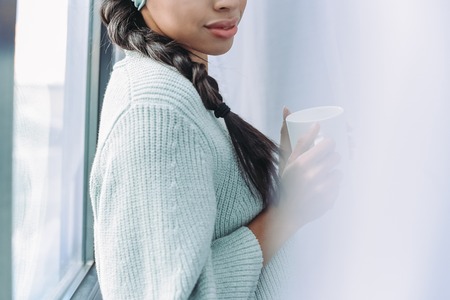 cropped image of mixed race girl in turquoise sweater holding cup of tea at homeの写真素材