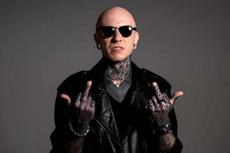 aggressive tattooed man in leather jacket and sunglasses showing middle fingers isolated on grey
