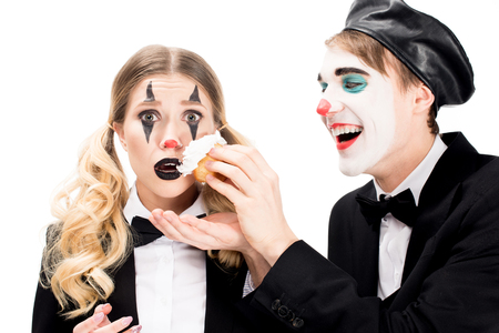 cheerful clown throwing cupcake in face of surprised woman isolated on whiteの写真素材