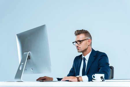 handsome businessman in suit and glasses looking at computer at table isolated on whiteの写真素材