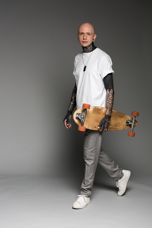 handsome tattooed man in white t-shirt walking with longboard and looking at camera on greyの写真素材