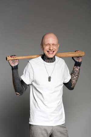 cheerful tattooed man with closed eyes standing with baseball bat on shoulders and laughing on greyの写真素材