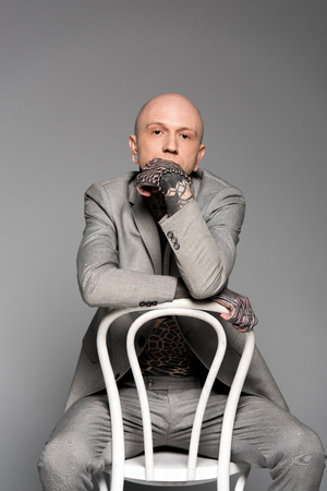 thoughtful tattooed man in suit sitting with hand on chin and looking at camera isolated on greyの写真素材