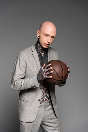 handsome bald tattooed man in suit holding basketball ball and looking at camera isolated on greyの写真素材