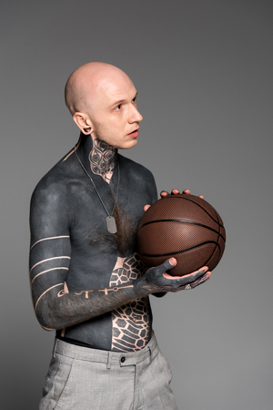 shirtless bald tattooed man holding basketball ball and looking away isolated on greyの写真素材