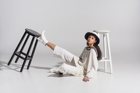 attractive african american young woman in stylish white clothes and hat sitting near black and white chairs on whiteの写真素材