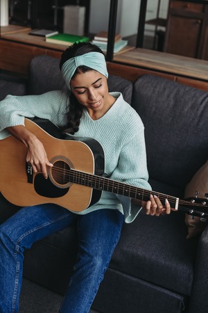 high angle view of beautiful mixed race girl in turquoise sweater and headband playing acoustic guitar on sofa at homeの写真素材