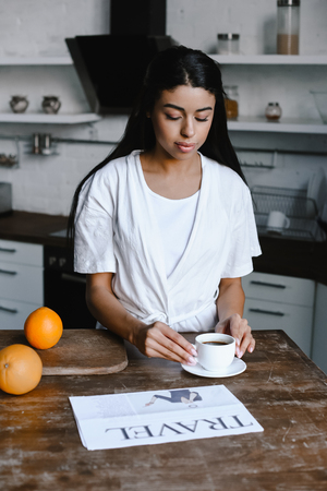 beautiful mixed race girl in white robe taking cup of coffee in morning in kitchenの写真素材
