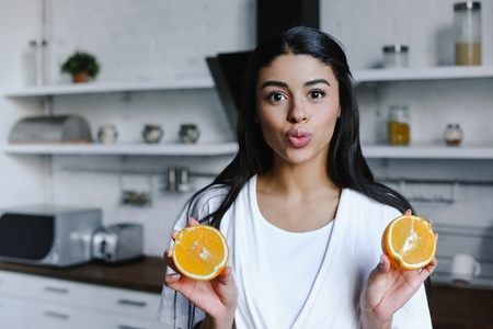 beautiful mixed race girl in white robe holding cut orange, grimacing and looking at camera in morning in kitchenの写真素材