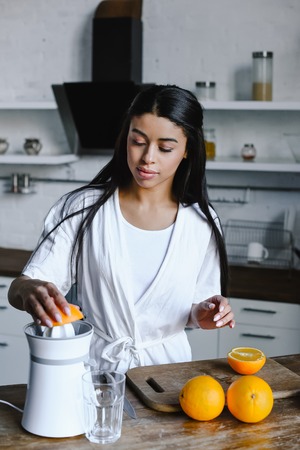 beautiful mixed race girl in white robe preparing orange juice in morning in kitchenの写真素材