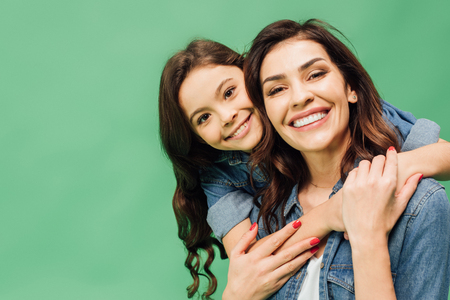 portrait of cheerful mother and daughter embracing and looking at camera isolated on greenの写真素材