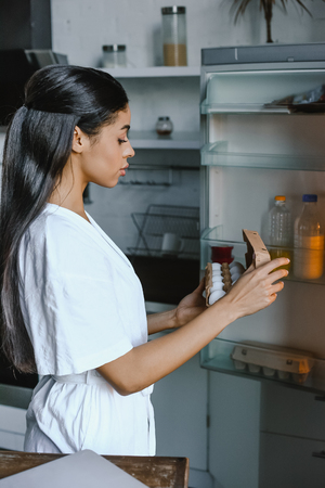 side view of beautiful mixed race girl in white robe taking tray with eggs from open fridge in morning in kitchenの写真素材