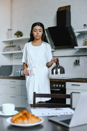 beautiful mixed race girl in white robe holding kettle in morning in kitchenの写真素材