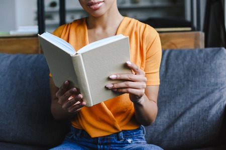 cropped image of mixed race girl in orange shirt reading book in living room
