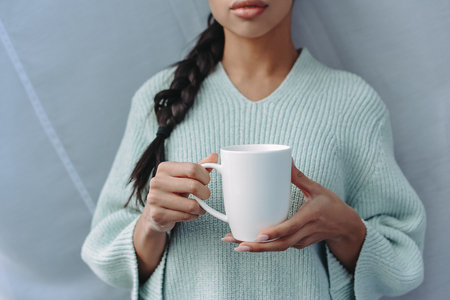 cropped image of mixed race girl in turquoise sweater holding cup of tea at homeの写真素材