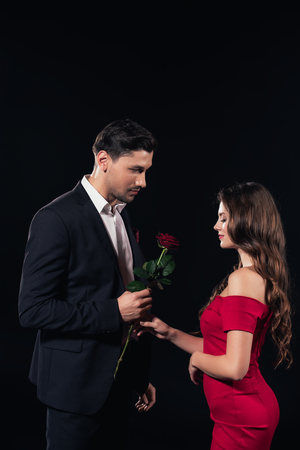 handsome man presenting red rose to woman in red dress isolated on blackの写真素材