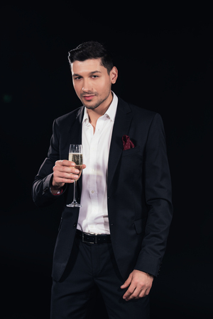 handsome man looking at camera and holding glass of champagne isolated on blackの写真素材