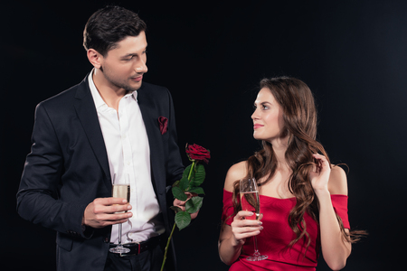 beautiful couple looking at each other, holding red rose and champagne glasses isolated on blackの写真素材