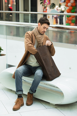 handsome man sitting on bench and holding bag in shopping mallの写真素材