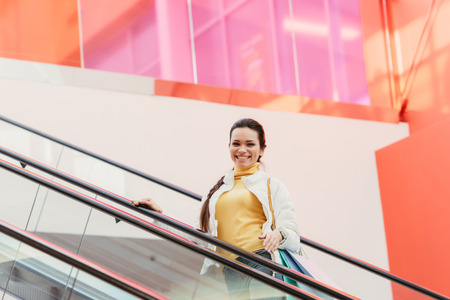 attractive girl with shopping bags smiling and looking at camera on escalatorの写真素材
