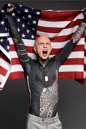 excited shirtless bare-chested man with tattoos holding american flag and screaming isolated on greyの写真素材