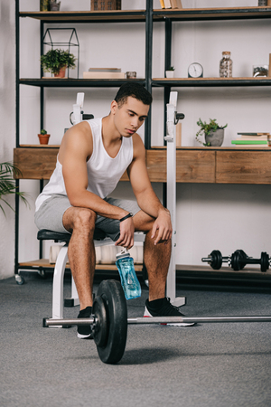 thoughtful bi-racial man sitting near barbell and holding sport bottleの写真素材