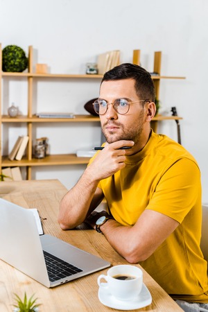 thoughtful man sitting in glasses near laptop and cup of coffee