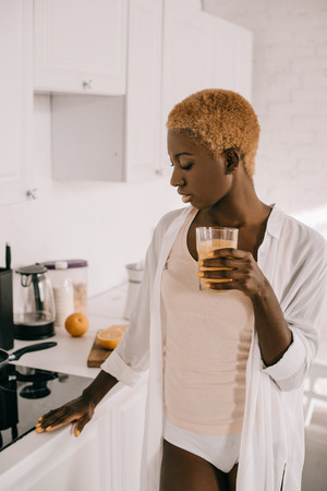 thoughtful african american woman holding glass of orange juice in kitchenの写真素材
