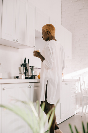 african american woman with short hair holding glass of orange juice in white kitchenの写真素材